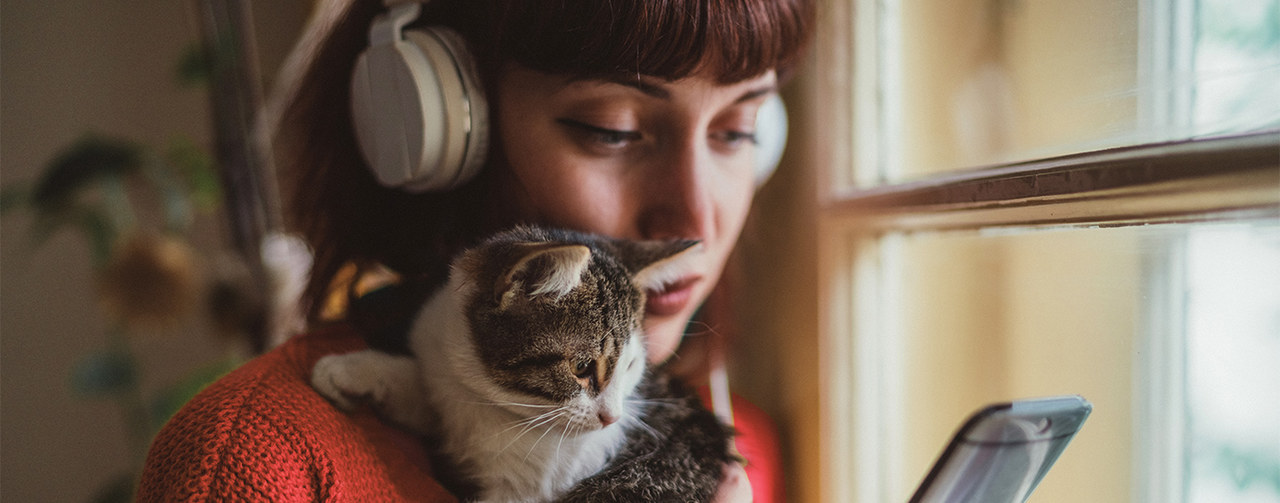Close-up of a young brunette woman with bangs and headphones, holding a cat and looking at her phone, near a window