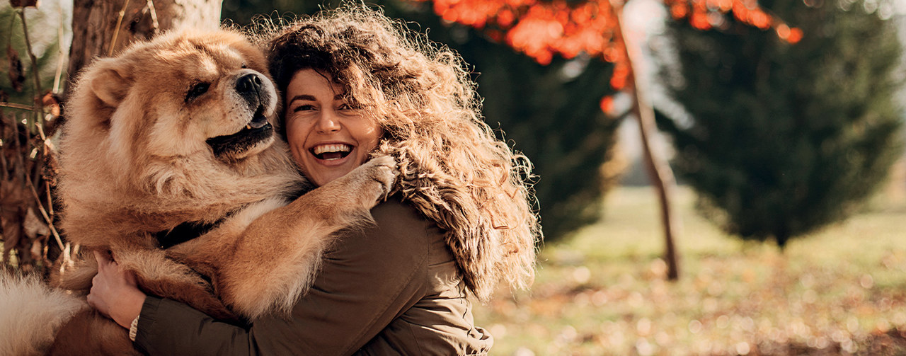 Symrise Pet Food's key image features a young woman with long, curly dark hair holding a large, hairy dog, a Chow-chow, who stands against an autumnal background