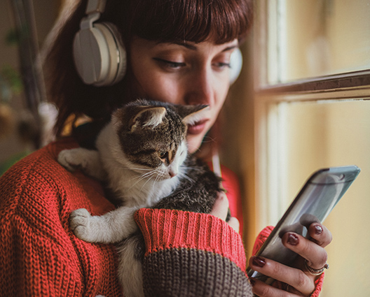 Close-up of a young brunette woman with bangs and headphones, holding a cat and looking at her phone, near a window